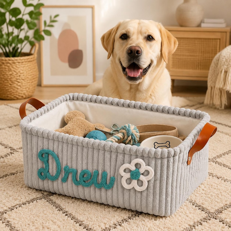 May include: A rectangular light gray corduroy storage basket with a white fabric lining and brown leather handles. The exterior features the name Drew in teal cursive yarn lettering and a white flower applique with a teal center.
