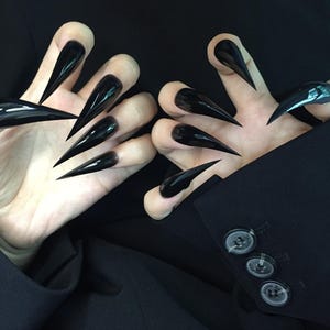 May include: Two hands with long, glossy black stiletto nails. The nails are sharply pointed and extend beyond the fingertips. The hands are positioned against a dark background, with a black blazer visible.