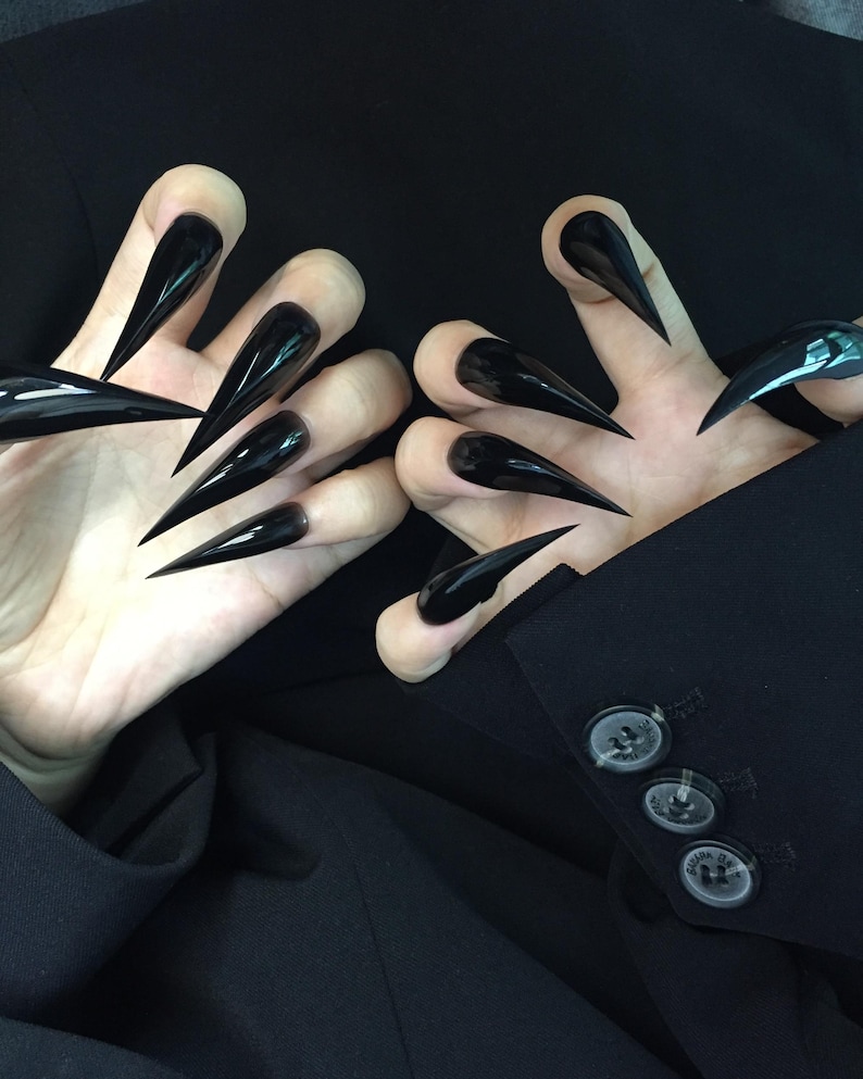 May include: Two hands with long, glossy black stiletto nails. The nails are sharply pointed and extend beyond the fingertips. The hands are positioned against a dark background, with a black blazer visible.