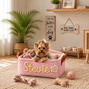 May include: A rectangular pink corduroy storage basket featuring the name Stevie in yellow script lettering and a white flower applique. The basket has brown leather handles and contains a small Yorkshire Terrier and a plush teddy bear toy.