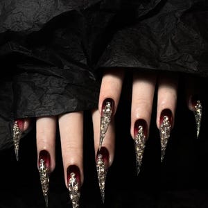 Puede incluir: Primer plano de u&ntilde;as largas y puntiagudas con un degradado de rojo oscuro a negro. Cada u&ntilde;a est&aacute; adornada con adornos plateados y ornamentados. Las u&ntilde;as est&aacute;n sobre un fondo oscuro y texturizado.