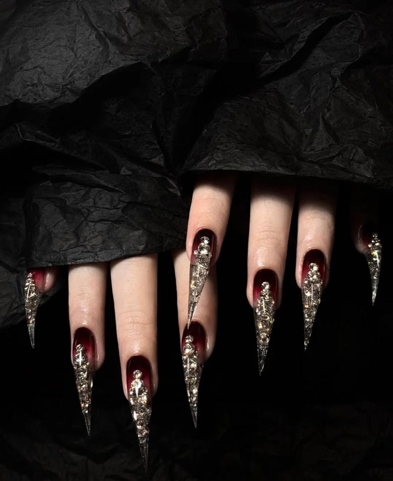 Puede incluir: Primer plano de u&ntilde;as largas y puntiagudas con un degradado de rojo oscuro a negro. Cada u&ntilde;a est&aacute; adornada con adornos plateados y ornamentados. Las u&ntilde;as est&aacute;n sobre un fondo oscuro y texturizado.