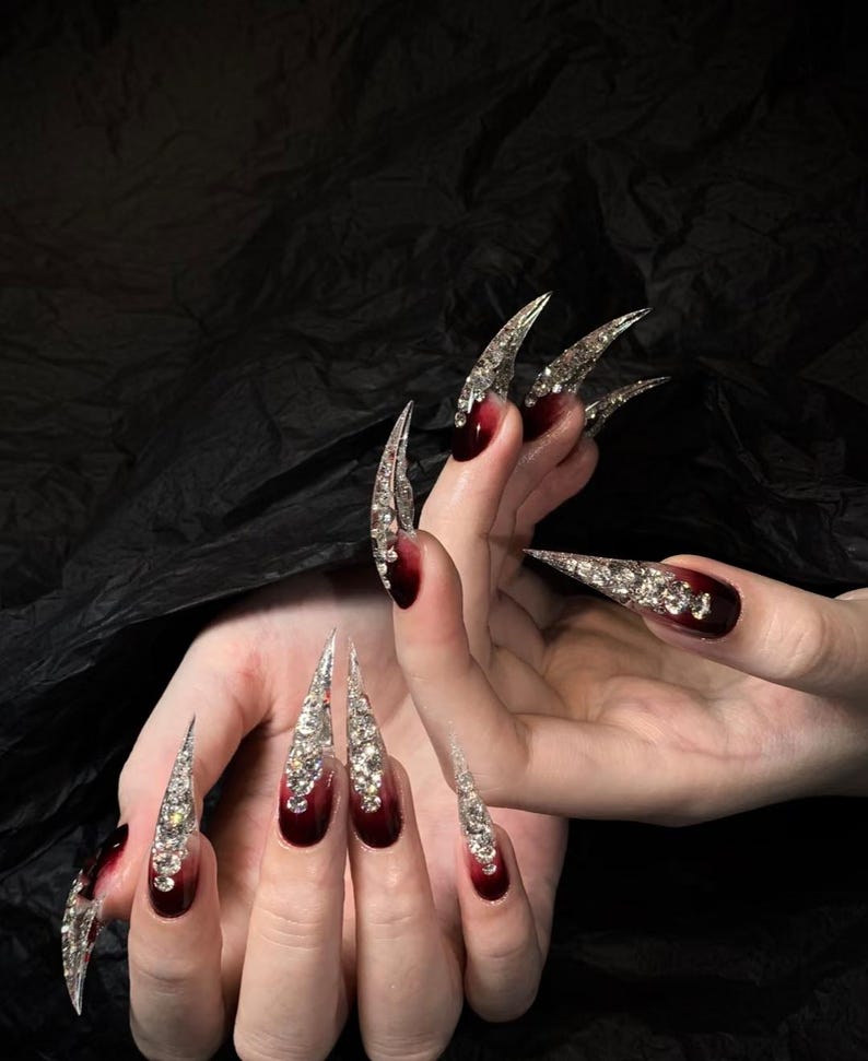 Puede incluir: U&ntilde;as artificiales largas y puntiagudas con un degradado de rojo oscuro a transparente, muy adornadas con pedrer&iacute;a plateada. Las u&ntilde;as son un accesorio dram&aacute;tico y glamuroso, perfecto para una ocasi&oacute;n especial o una declaraci&oacute;n de moda audaz.