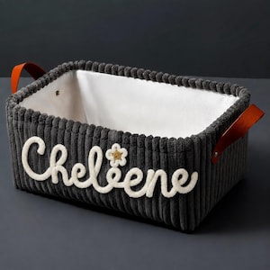 May include: A rectangular dark gray corduroy storage basket with a white fabric interior lining. The exterior features the white embroidered name Cheleene with a small flower icon. Two brown leather handles are attached with silver metal rivets.