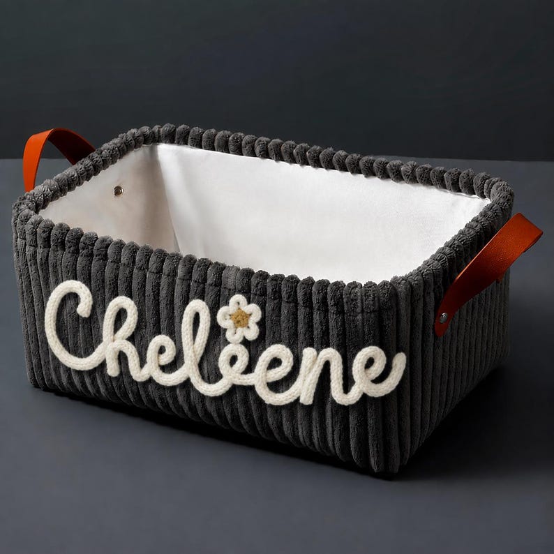 May include: A rectangular dark gray corduroy storage basket with a white fabric interior lining. The exterior features the white embroidered name Cheleene with a small flower icon. Two brown leather handles are attached with silver metal rivets.