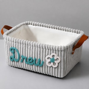 May include: A rectangular storage basket made of grey corduroy fabric with a white interior lining. The exterior features the name Drew in teal yarn lettering and a white crocheted flower with a teal center. It has brown faux leather handles.