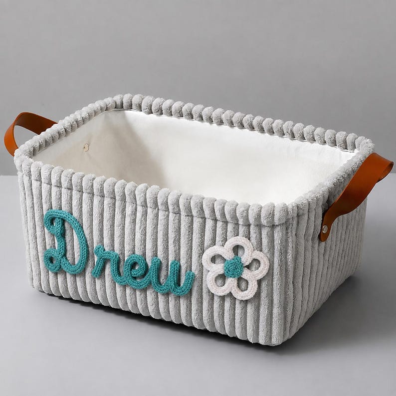 May include: A rectangular storage basket made of grey corduroy fabric with a white interior lining. The exterior features the name Drew in teal yarn lettering and a white crocheted flower with a teal center. It has brown faux leather handles.