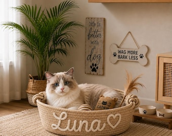 May include: A round, beige woven rope pet bed featuring the name Luna and a heart shape in white rope lettering on the front. A fluffy cat sits inside the bed alongside a small brown catnip toy and a feather wand toy.