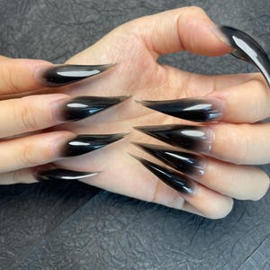 May include: A hand displaying long, pointed acrylic nails. The nails transition from a clear base to a gradient of black, creating a dramatic and stylish effect. The nails are sharply pointed, adding to the bold aesthetic.