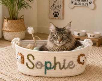 May include: A round, off-white woven cotton rope basket featuring the name Sophie in green and grey yarn lettering. The basket is decorated with two small, plush lion faces on the sides and has knotted rope handles on each end.