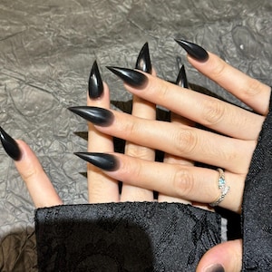 May include: Black stiletto nails with a glossy finish. The nails are long and sharply pointed, creating a dramatic effect. A silver ring with a small blue stone is visible on one finger.