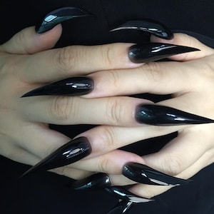 May include: A close-up shot of a set of long, glossy black stiletto nails. The nails are sharply pointed and reflect light, creating a mirror-like effect. The manicure is a bold statement, showcasing a sleek and modern aesthetic.