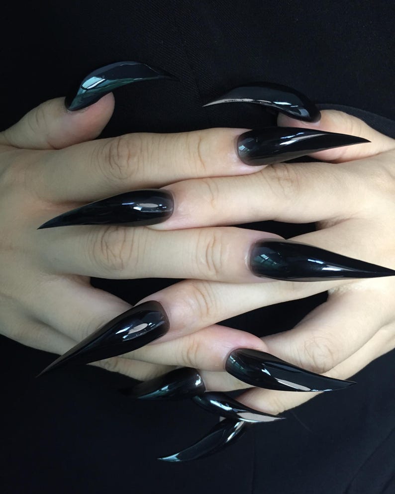 May include: A close-up shot of a set of long, glossy black stiletto nails. The nails are sharply pointed and reflect light, creating a mirror-like effect. The manicure is a bold statement, showcasing a sleek and modern aesthetic.
