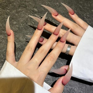 May include: A set of long, pointed, translucent nails with a gradient effect. The nails transition from a dark red at the base to a pale, almost clear tip. The nails are a dramatic, fashionable accessory.
