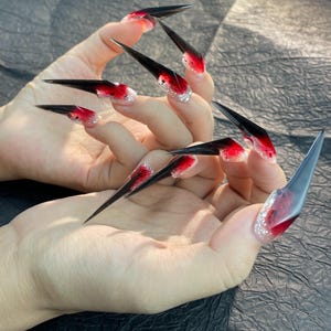 May include: A set of long, pointed acrylic nails. Each nail features a gradient design, transitioning from a red base to a black tip. The base of each nail is adorned with silver glitter.