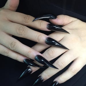 May include: Close-up of hands with long, glossy, black stiletto nails. The nails are sharply pointed and reflect light, creating a sleek and dramatic appearance. The nails are a solid, opaque black colour.