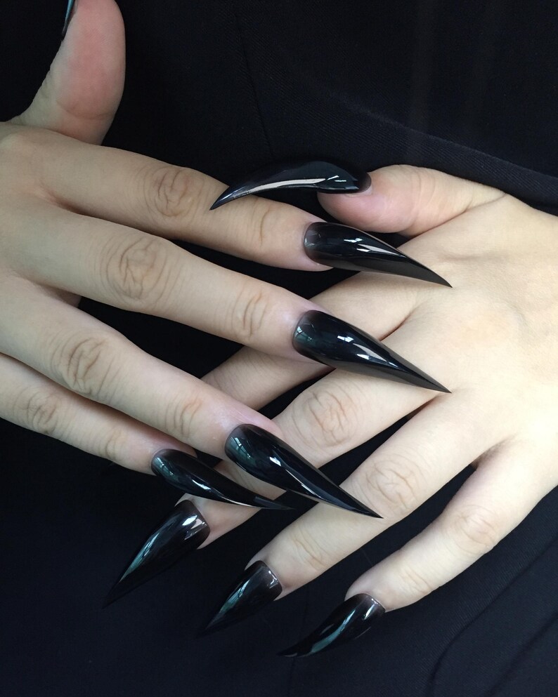 May include: Close-up of hands with long, glossy, black stiletto nails. The nails are sharply pointed and reflect light, creating a sleek and dramatic appearance. The nails are a solid, opaque black colour.