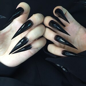 May include: A close-up shot of two hands displaying long, glossy black stiletto nails. The nails are sharply pointed and reflect light, creating a dramatic effect. The hands are set against a dark background, emphasising the nails' bold appearance.