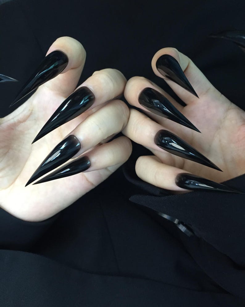 May include: A close-up shot of two hands displaying long, glossy black stiletto nails. The nails are sharply pointed and reflect light, creating a dramatic effect. The hands are set against a dark background, emphasising the nails' bold appearance.