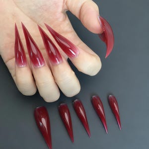 Puede incluir: Un conjunto de uñas postizas largas y puntiagudas, de color burdeos. Las uñas son de un rojo intenso en las puntas, desvaneciéndose a un tono más claro cerca de la base. También se muestran varias uñas individuales, mostrando el diseño y el color.