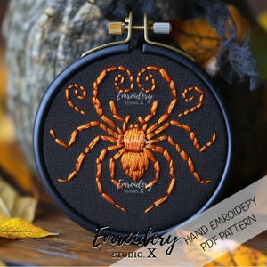 Puede incluir: Un bastidor de bordado negro con un diseño de araña naranja. La araña está bordada con detalles, y el texto "Embroidery Studio X" es visible. El bastidor se encuentra sobre un fondo borroso con hojas de otoño.