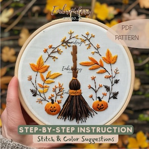 May include: An embroidery hoop containing white fabric with a hand-stitched design of a central brown broomstick flanked by two orange jack-o'-lanterns and autumn foliage in shades of yellow and orange. The text reads PDF PATTERN.