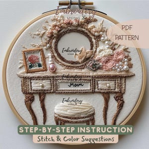 May include: An embroidered design featuring a vanity table with a mirror, flowers, and a stool. The design includes the word "Mom" on the drawer and the text "Embroidery Studio". The design is in a wooden embroidery hoop.