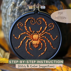Puede incluir: Un bastidor de bordado negro que contiene un diseño de araña naranja cosido a mano sobre tela negra. La araña presenta un trabajo de hilo detallado con patas curvadas. El texto incluye Embroidery Studio X, PDF PATTERN y STEP-BY-STEP INSTRUCTION.