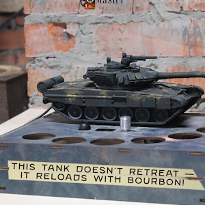 以下が含まれることがあります： 長い砲身と回転砲塔を備えた濃い緑色の玩具戦車が木製の台座に置かれています。 台座にはいくつかの円形の切り欠きがあり、「この戦車は後退しません - バーボンでリロードします！」と書かれた看板があります。 「Pouring Master」のロゴが見えます。