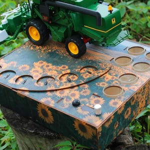 Könnte beinhalten: Ein grüner Spielzeug-Mähdrescher mit gelben Rädern steht auf einer rechteckigen Holzbox mit Sonnenblumenmuster. Die Box verfügt über mehrere kreisförmige Aussparungen und einen kleinen silbernen Knopf auf der Oberfläche.