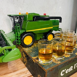 Puede incluir: Una cosechadora de juguete verde con ruedas amarillas y la palabra "HARVESTER" impresa en el lateral. Varios vasos de chupito llenos de líquido ámbar están colocados sobre una caja decorativa con detalles florales y texto.