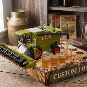 Peut inclure: Un modèle réduit de moissonneuse-batteuse Claas vert et blanc posé sur un socle en bois avec le texte CUSTOM LOGO. La moissonneuse possède un tuyau de décharge noir versant un liquide ambré dans quatre verres à shot transparents sur le socle.