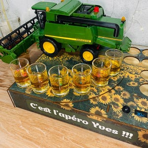 Könnte beinhalten: Ein grüner und gelber Spielzeug-Mähdrescher steht hinter einem hölzernen Servierbrett mit Sonnenblumenmuster. Das Brett enthält sechs klare Schnapsgläser, die mit einer goldenen Flüssigkeit gefüllt sind, und den Aufdruck C'est l'apéro Yves !!!