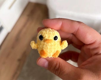 May include: A small, handmade yellow crochet chick with orange feet and beak, and large black eyes. The chick has small wings and is held in a person's hand. The background is blurred.