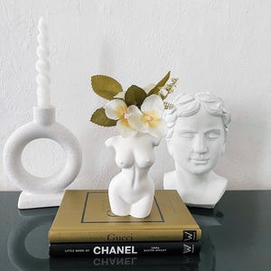 Könnte beinhalten: Weiße Keramikskulptur eines weiblichen Torsos mit einem Strauß weißer und gelber Blumen im Busen. Die Skulptur steht auf einer schwarzen Oberfläche mit zwei goldenen Büchern mit den Titeln "Little Book of Gucci" und "Little Book of Chanel".