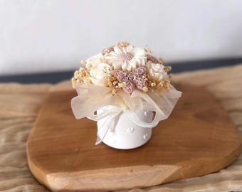 May include: A white cylindrical candle container decorated with small embossed hearts, topped with a cluster of cream and dusty pink floral-shaped wax pieces, dried baby's breath, and a translucent white ribbon tied in a bow around the base.