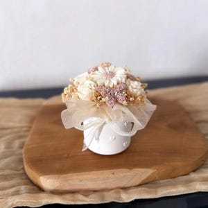 Könnte beinhalten: Eine kleine, weiße Keramikvase mit einem Strauß künstlicher Blumen. Das Arrangement umfasst weiße und rosa Blüten, mit einem transparenten Stoff und einem Band. Die Vase steht auf einem Holzbrett.