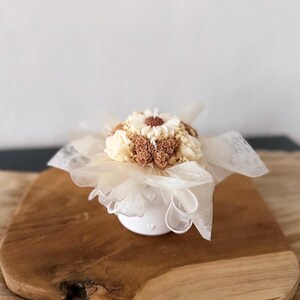 May include: A white ceramic candle holder with a floral arrangement of cream and brown wax flowers. The candle is wrapped in delicate white fabric and sits on a wooden board.