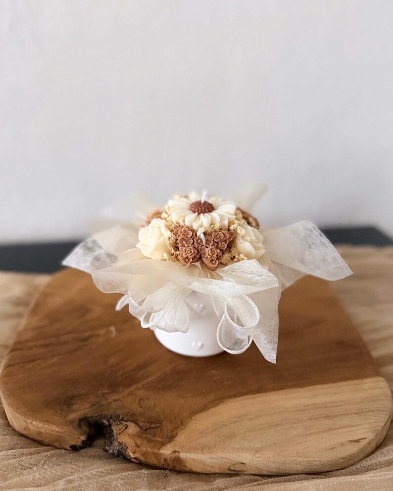 May include: A white ceramic candle holder with a floral arrangement of cream and brown wax flowers. The candle is wrapped in delicate white fabric and sits on a wooden board.