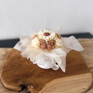 May include: A decorative candle shaped like a floral bouquet. The candle features a large daisy in the center, surrounded by smaller flowers in shades of cream and brown. It sits in a small white vase and is wrapped in sheer fabric.