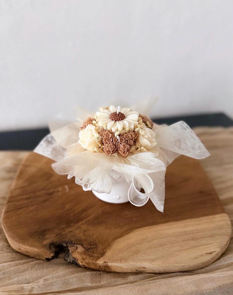 May include: A decorative candle shaped like a floral bouquet. The candle features a large daisy in the center, surrounded by smaller flowers in shades of cream and brown. It sits in a small white vase and is wrapped in sheer fabric.