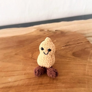 Könnte beinhalten: Eine handgefertigte, beige Häkel-Erdnuss-Puppe mit einem lächelnden Gesicht und braunen Füßen. Die Puppe sitzt auf einer Holzoberfläche. Die Erdnuss-Puppe ist ein einzigartiger, skurriler Artikel.