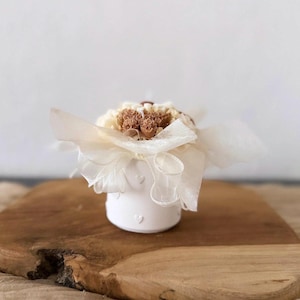 May include: A small, white ceramic container with heart-shaped cutouts, holding a bouquet of dried flowers. The arrangement is wrapped in cream-colored paper and tied with a sheer ribbon. The container sits on a wooden surface.