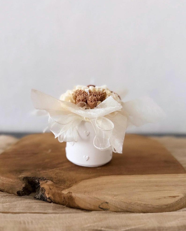 May include: A small, white ceramic container with heart-shaped cutouts, holding a bouquet of dried flowers. The arrangement is wrapped in cream-colored paper and tied with a sheer ribbon. The container sits on a wooden surface.