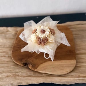 May include: A decorative candle shaped like a bouquet of flowers, featuring a large daisy and smaller blossoms in shades of cream and brown. The candle is wrapped in delicate white fabric and sits on a wooden board.