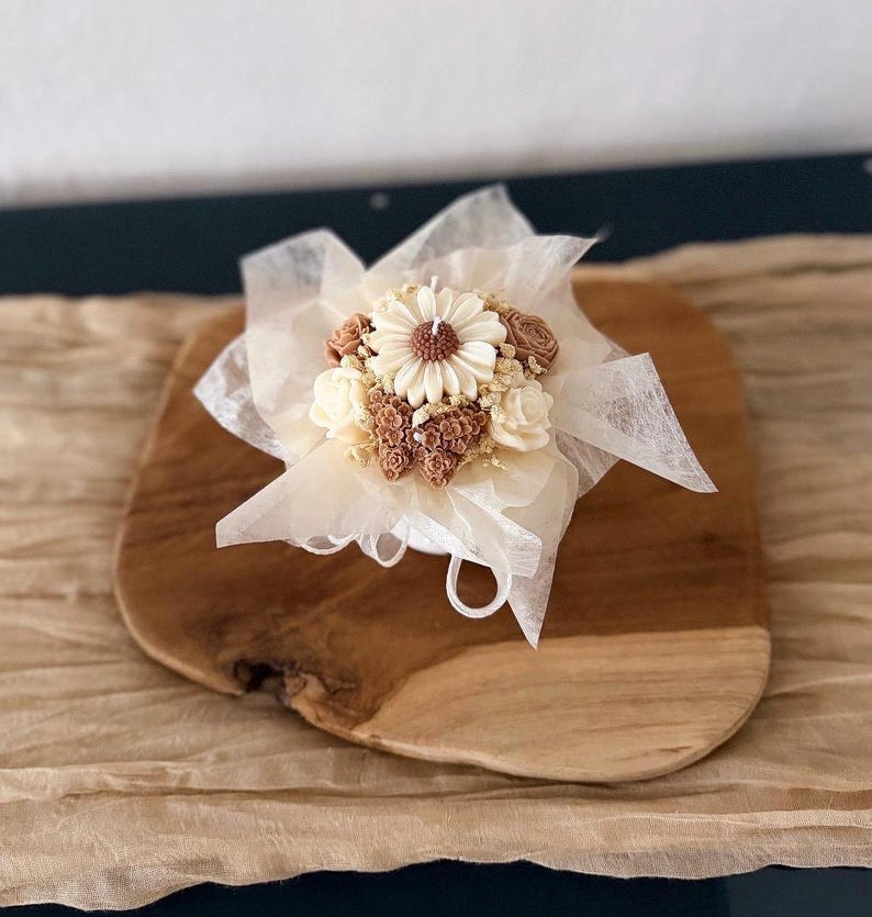May include: A decorative candle shaped like a bouquet of flowers, featuring a large daisy and smaller blossoms in shades of cream and brown. The candle is wrapped in delicate white fabric and sits on a wooden board.