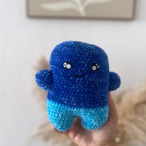 May include: A blue and light blue crocheted plush toy with a smiling face and large eyes. The toy is shaped like a person with arms and legs.