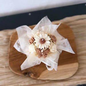 May include: A decorative candle shaped like a bouquet of flowers, featuring a large daisy at the center, surrounded by smaller flowers in shades of cream and brown. The candle is wrapped in delicate white fabric and tied with a ribbon, presented on a wooden surface.