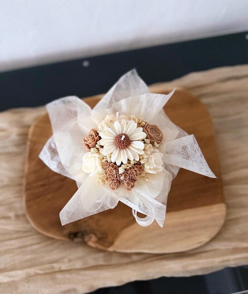 May include: A decorative candle shaped like a bouquet of flowers, featuring a large daisy at the center, surrounded by smaller flowers in shades of cream and brown. The candle is wrapped in delicate white fabric and tied with a ribbon, presented on a wooden surface.