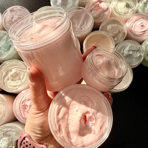 Può includere: Una collezione di barattoli in plastica trasparente riempiti con sostanze dense e montate, simili a una crema, in varie tonalità pastello, tra cui rosa chiaro, bianco e verde pallido. I barattoli hanno bordi lisci e arrotondati.